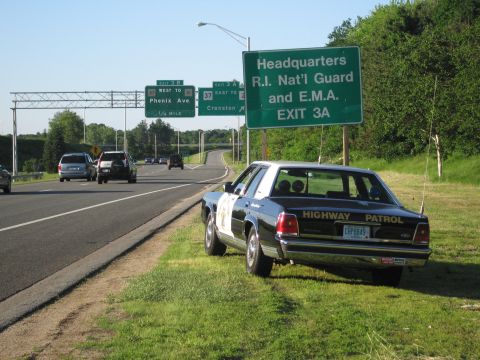 1991 Ford LTD Crown Victoria - black-and-white HIGHWAY PATROL sedan sits on the grass below a green highway sign for R.I. Nat'l Guard Headquarters, Exit 3A.