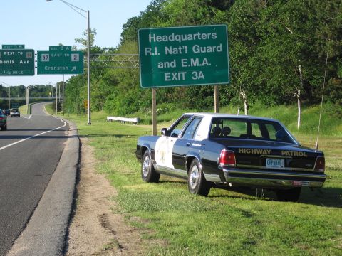 1991 Ford LTD Crown Victoria - black-and-white HIGHWAY PATROL sedan with a gold star door shield rests on the grass beneath a green R.I. Nat'l Guard exit sign.