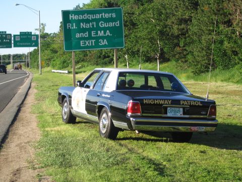 1991 Ford LTD Crown Victoria - black-and-white HIGHWAY PATROL sedan with a star door emblem and CHP8849 plate sits on the grass below a green highway exit sign.