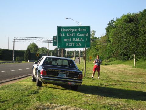 1991 Ford LTD Crown Victoria - black-and-white HIGHWAY PATROL sedan sits on the highway shoulder while a man in a gray shirt and red shorts stands beside the exit sign.