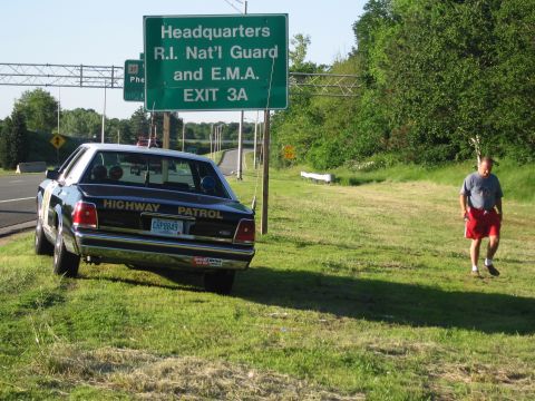 1991 Ford LTD Crown Victoria - black-and-white HIGHWAY PATROL Ford sedan is parked under a green exit sign as a man in red shorts walks across the grass.