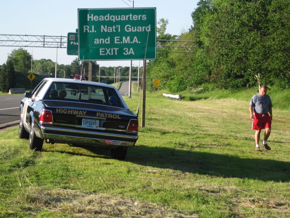 Black-and-white Highway Patrol Ford sedan with a man in red shorts walking on the grass
