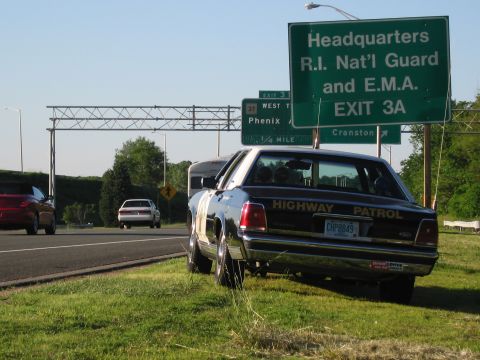 1991 Ford LTD Crown Victoria - rear of a black-and-white HIGHWAY PATROL sedan with CHP8849 plate sits on the highway shoulder beneath green guide signs.