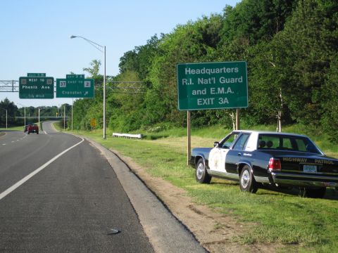 1991 Ford LTD Crown Victoria - black-and-white HIGHWAY PATROL sedan with a gold star door shield sits on the grass shoulder of a curving highway near a green exit sign.