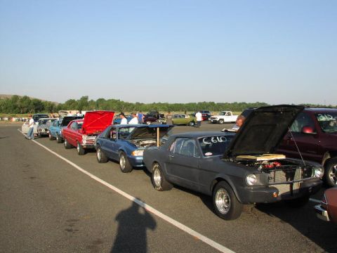 1981 Ford Bronco - Several cars are lined up at a drag strip, with one dark gray coupe prominently displayed with its hood open.