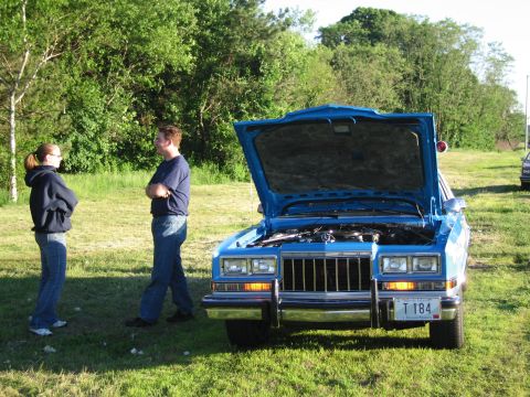 1991 Ford LTD Crown Victoria - Two people stand talking beside a blue sedan with its hood raised in a grassy field at sunset, the engine bay exposed.
