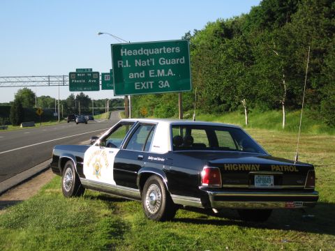 1991 Ford LTD Crown Victoria - black-and-white HIGHWAY PATROL sedan with gold star shield and CHP8849 plate rests on the grass below a green R.I. exit sign.