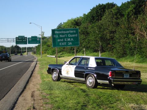 1991 Ford LTD Crown Victoria - black-and-white HIGHWAY PATROL sedan with a gold star door shield sits on the grassy shoulder of a highway beneath green guide signs.