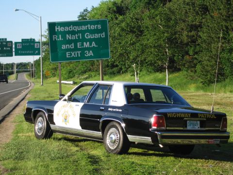 1991 Ford LTD Crown Victoria - black-and-white HIGHWAY PATROL sedan with a gold star door shield and CHP8849 plate is parked on grass next to a green exit sign.