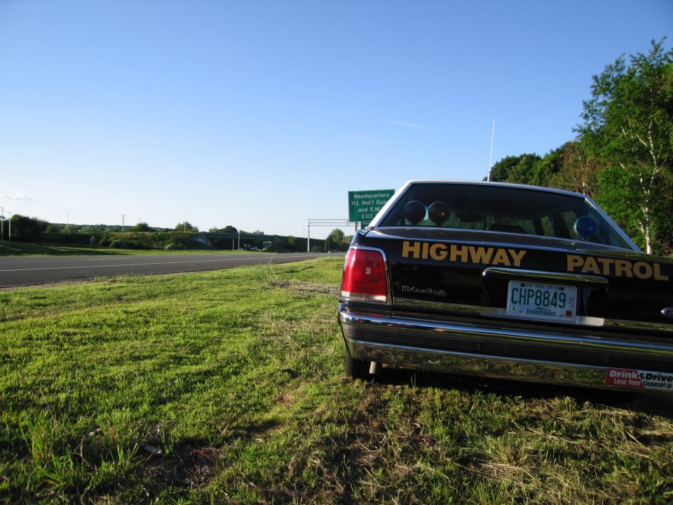 Rear of a black Highway Patrol sedan with roof lights parked on a grassy shoulder