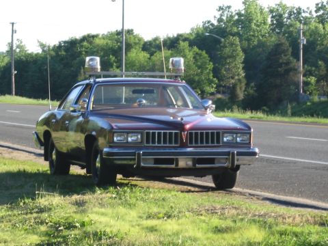 1991 Ford LTD Crown Victoria - maroon two-door coupe fitted with roof emergency lights and twin spotlights drives along a rural roadside lined with trees.