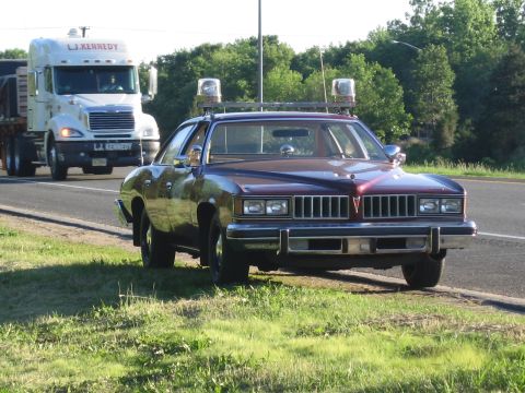 1991 Ford LTD Crown Victoria - maroon Pontiac coupe with roof emergency lights and spotlights drives down a rural road as an LJ Kennedy semi truck approaches.