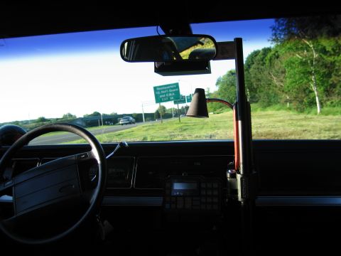 1991 Ford LTD Crown Victoria - Looking out an older sedan's windshield past the wheel and radio gear toward a highway exit sign and grassy roadside.
