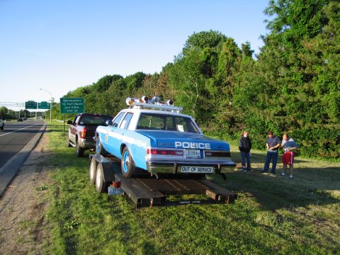 1991 Ford LTD Crown Victoria - blue-and-white POLICE car marked OUT OF SERVICE sits on a flatbed trailer by a roadside as several people stand nearby.