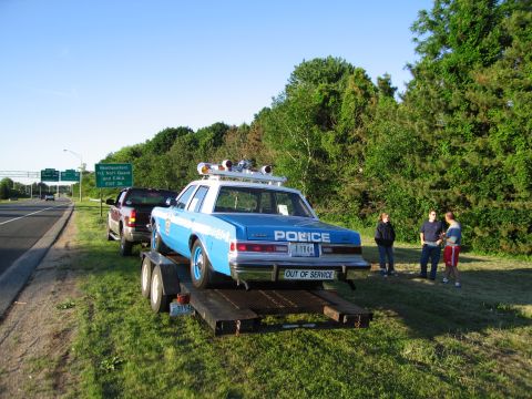 1991 Ford LTD Crown Victoria - blue-and-white police sedan loaded on a flatbed trailer near a wooded roadside, with several onlookers standing beside it.