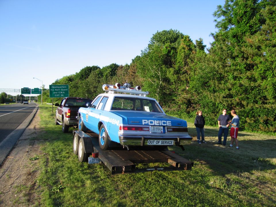 Police car on flatbed trailer with people standing by green roadside