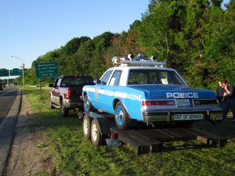 1991 Ford LTD Crown Victoria - blue POLICE sedan marked OUT OF SERVICE rides a flatbed trailer pulled by a red pickup along a grassy highway shoulder.