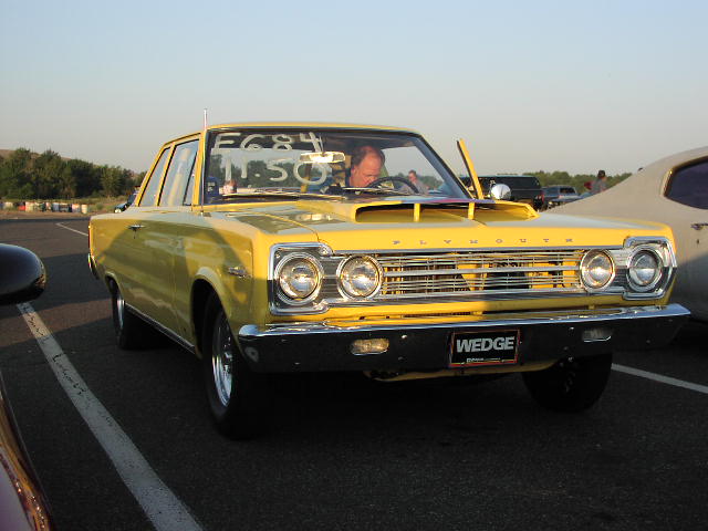 Yellow Plymouth Satellite with a hood scoop and a driver. Another car is parked next to it