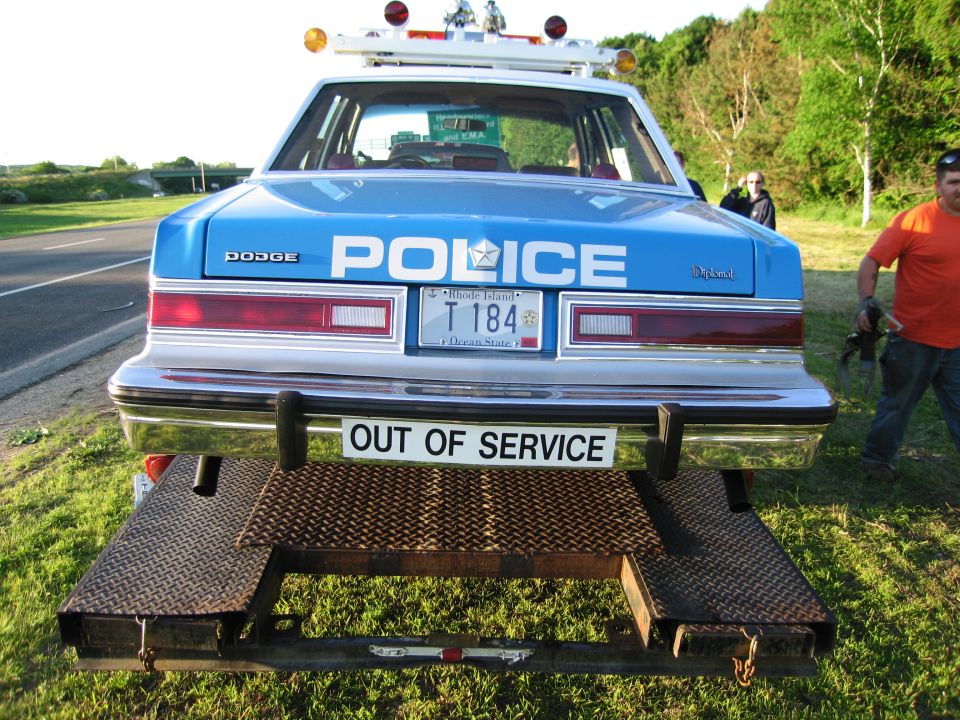 Rear of blue Dodge POLICE car on trailer, man standing at right
