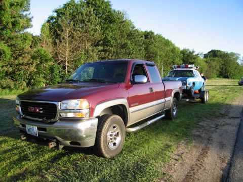 1991 Ford LTD Crown Victoria - maroon GMC extended-cab pickup parked on grass while towing a flatbed trailer carrying a blue police car.