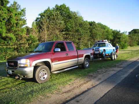 1991 Ford LTD Crown Victoria - maroon GMC pickup hitched to a flatbed trailer holding a blue police car, parked on grass beside a paved road.