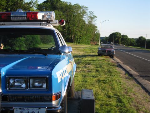 1991 Ford LTD Crown Victoria - Close front view of a blue POLICE car with roof lightbar loaded on a trailer, with a road and second vehicle in the distance.