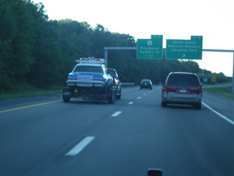1991 Ford LTD Crown Victoria - blue police car rides a flatbed trailer being towed along a multi-lane highway past green overhead exit signs.
