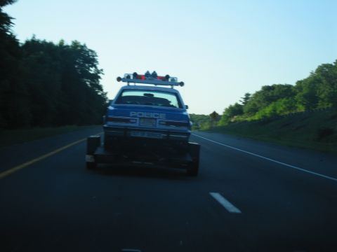 1991 Ford LTD Crown Victoria - Rear view of a blue POLICE car on a flatbed trailer being towed along a tree-lined two-lane road.