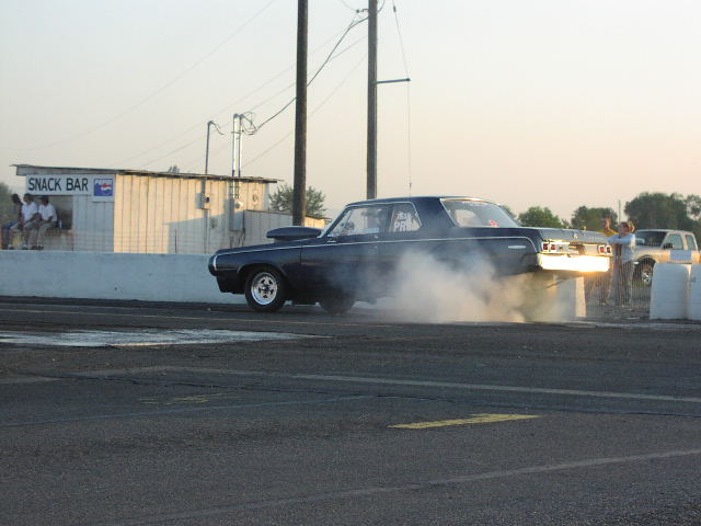 Dark drag car doing a burnout on a track, smoke billowing from tires. Snack bar in background