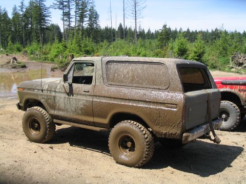 1979 Ford Bronco - Driver-side rear three-quarter view of a lifted full-size Ford Bronco caked head-to-toe in dried brown mud, parked on a gravel staging area…