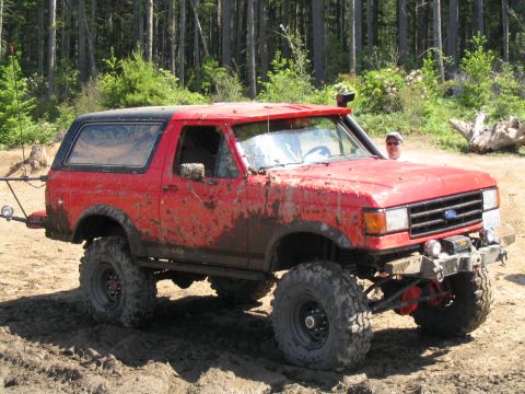 1979 Ford Bronco - Mud-splattered red full-size Ford Bronco with a black hardtop, photographed from the 3/4 front on a dirt trail cut through evergreen forest…