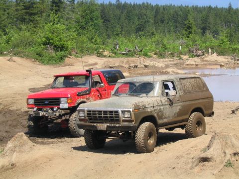 1979 Ford Bronco - Two off-road vehicles, a mud-caked 1978 Ford Bronco and a red Ford Bronco, are parked on a dirt track with trees in the background.