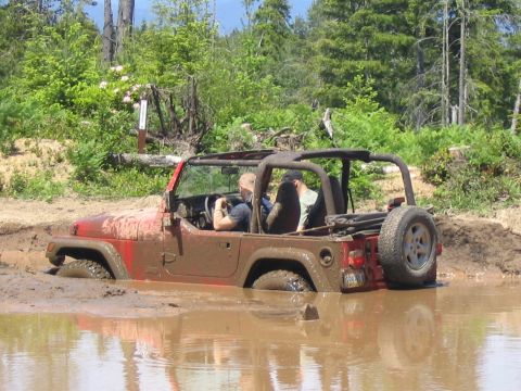 1979 Ford Bronco - A red Jeep is stuck in a muddy puddle off-road, with two occupants visible inside.