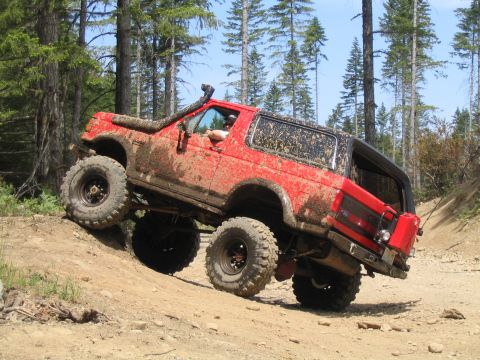 1979 Ford Bronco - A heavily modified red SUV navigates a steep, muddy incline on a wooded trail.