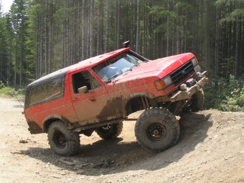 1979 Ford Bronco - Mud-splattered red full-size Ford Bronco flexing on a dirt berm at a forest trailhead, shown from the driver-side front three-quarter with…
