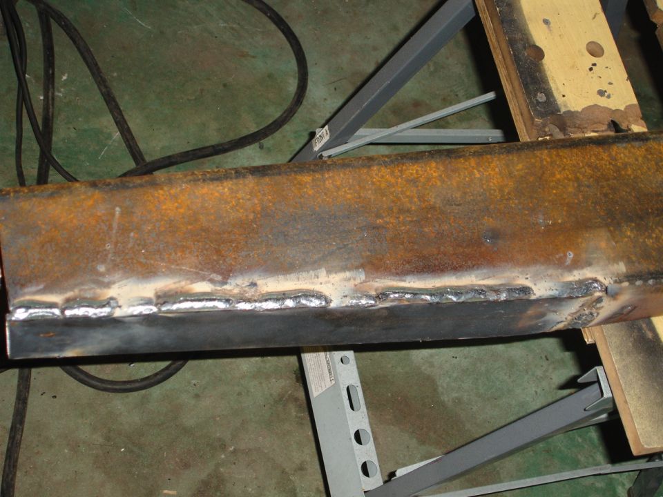 Close-up of a rusty metal beam with fresh welds, resting on a stand. A black cable is visible