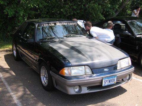1989 Ford Mustang - This 1989 Ford Mustang features a grey hood and aftermarket wheels, seen here at an outdoor gathering.