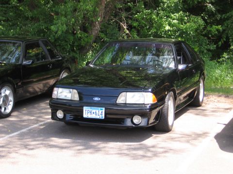 1989 Ford Mustang - A black coupe is parked in a lot with trees in the background. Another car is visible to the left.
