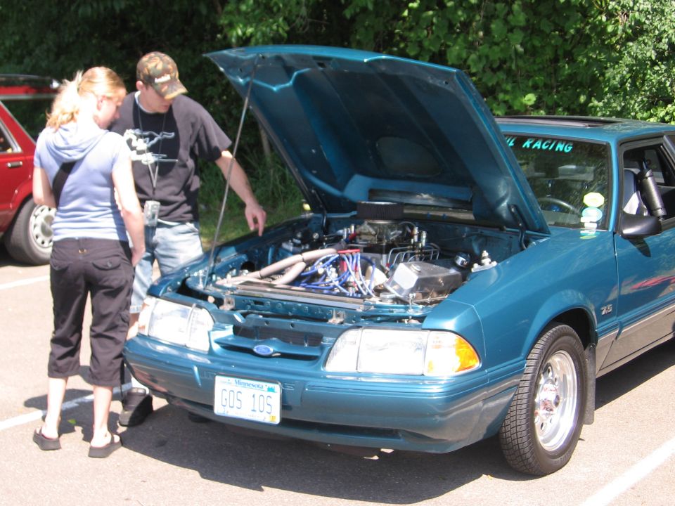 Teal 1989 Ford Mustang coupe with hood open, showing engine. Two people looking at it. Another red car is partially visible