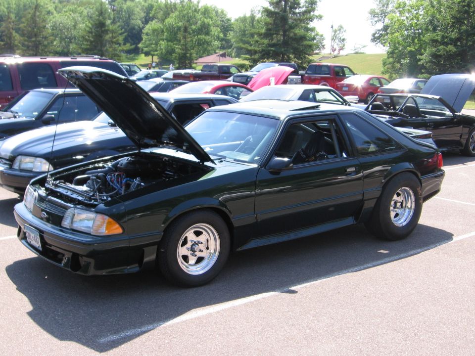 Dark green 1989 Ford Mustang parked with hood open, showing engine. Other cars visible in background