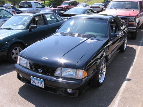 1989 Ford Mustang - A black Ford Mustang is parked in a lot with other vehicles. The car has chrome wheels and a blue license plate.