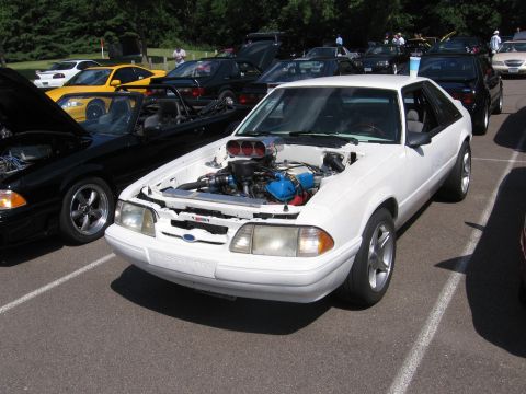 1989 Ford Mustang - A white Ford Mustang with its hood open, revealing a custom engine, parked among other vehicles.