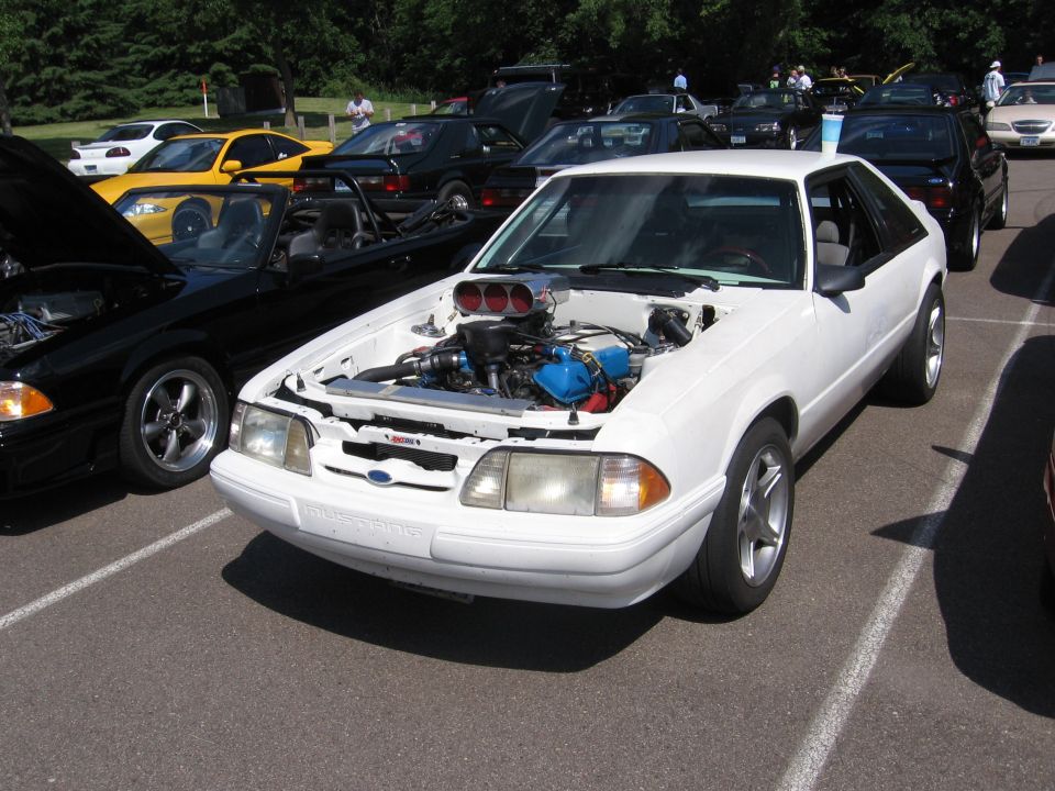 White Ford Mustang with hood open, showing a supercharger and blue engine components. Modified front end