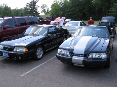 1989 Ford Mustang - Two Ford Mustangs are parked side-by-side in an outdoor lot with other vehicles and people.