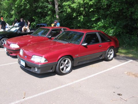 1989 Ford Mustang - Two red Ford Mustangs are parked side-by-side in an outdoor lot with trees in the background.