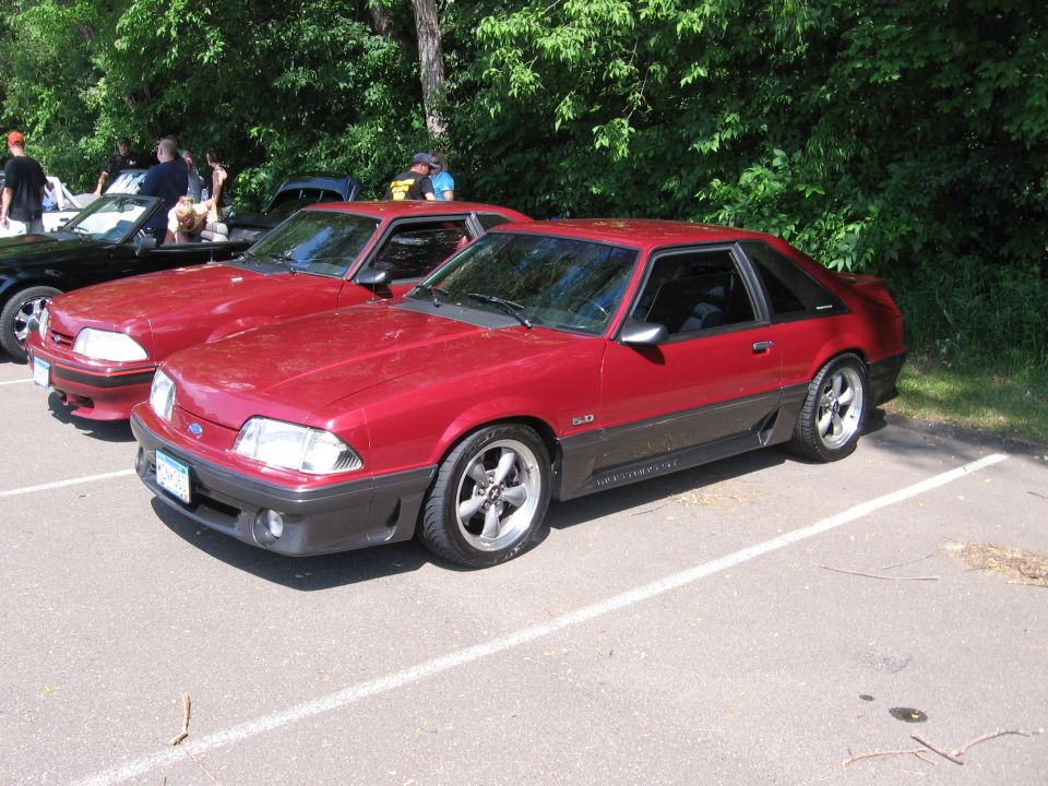 Red 1987 Ford Mustang coupe parked in a lot, showing its 3/4 front view with dark gray lower body cladding and aftermarket wheels