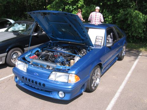 1989 Ford Mustang - A blue 1989 Ford Mustang is displayed with its hood up, revealing the engine. Another vehicle is parked nearby.