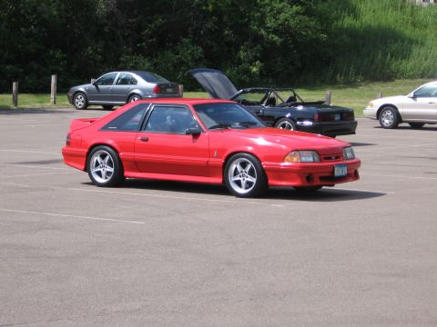 1989 Ford Mustang - A red coupe with a body kit and five-spoke wheels is parked in a lot with other vehicles.