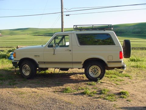 1988 Ford Bronco - A side view of a cream-colored Ford Bronco with a roof rack and spare tire, parked outdoors.