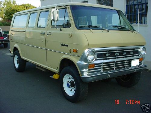 Tan Ford Econoline van with chrome grille and side windows. It has off-road tires and a yellow jack under the front passenger side